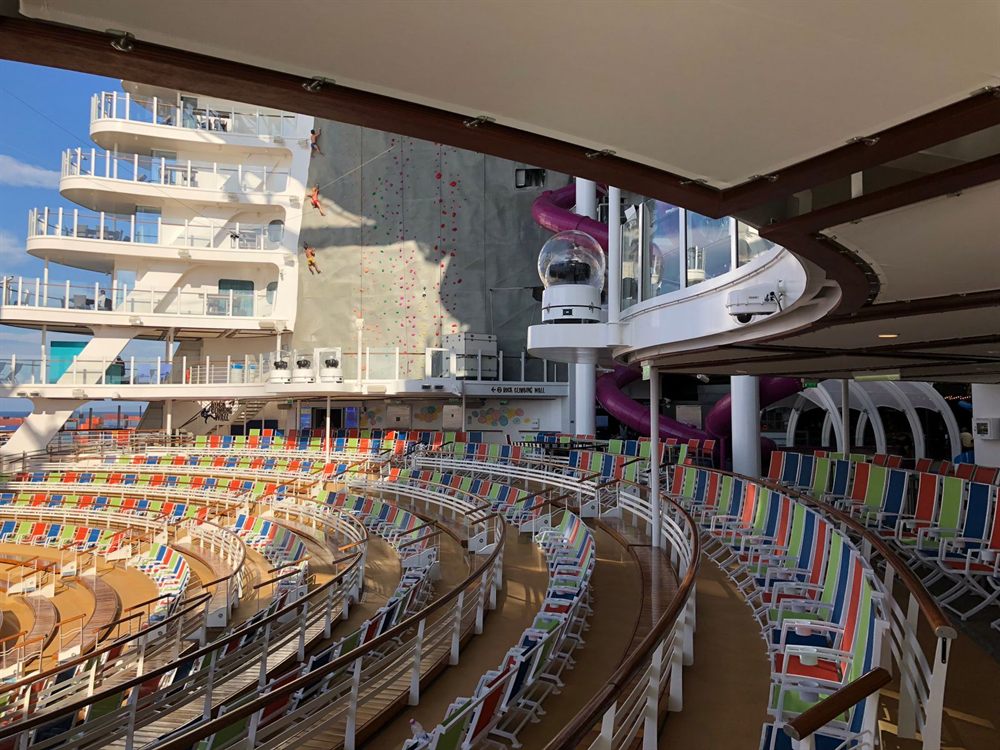 Rows of colorful seats around an open-air venue on a cruise ship, with a climbing wall behind