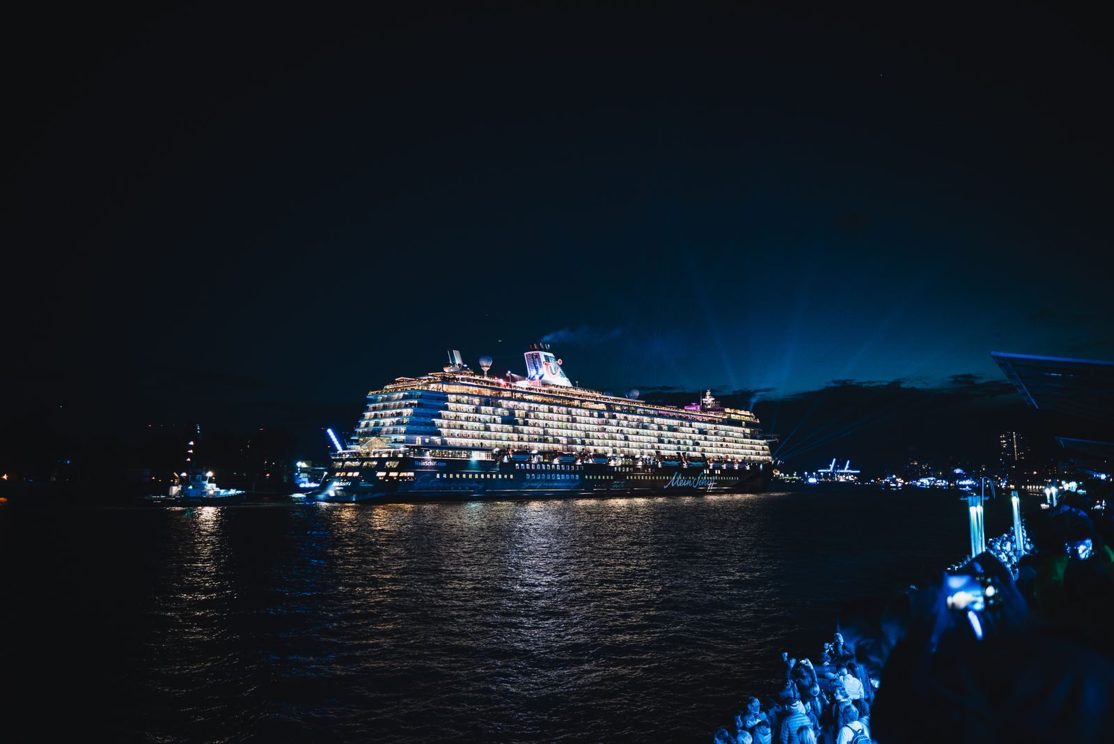 Giant cruise ship lit up at night as onlookers watch from the shore, like the world’s largest cruise ship in 2026 sailing in